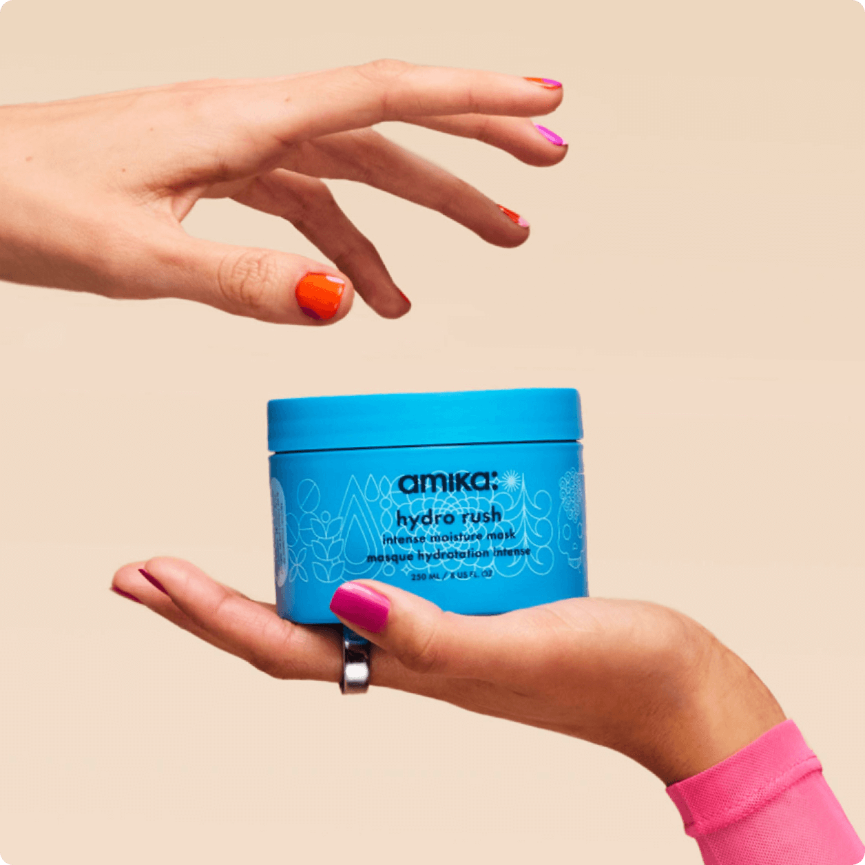 a hand holds amika's hydro rush intense moisture mask as a second hand above reaches for it against a cream background