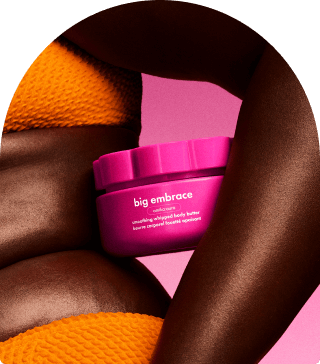 a model holds a container of big embrace body butter against their torso. they stand in front of a pink background