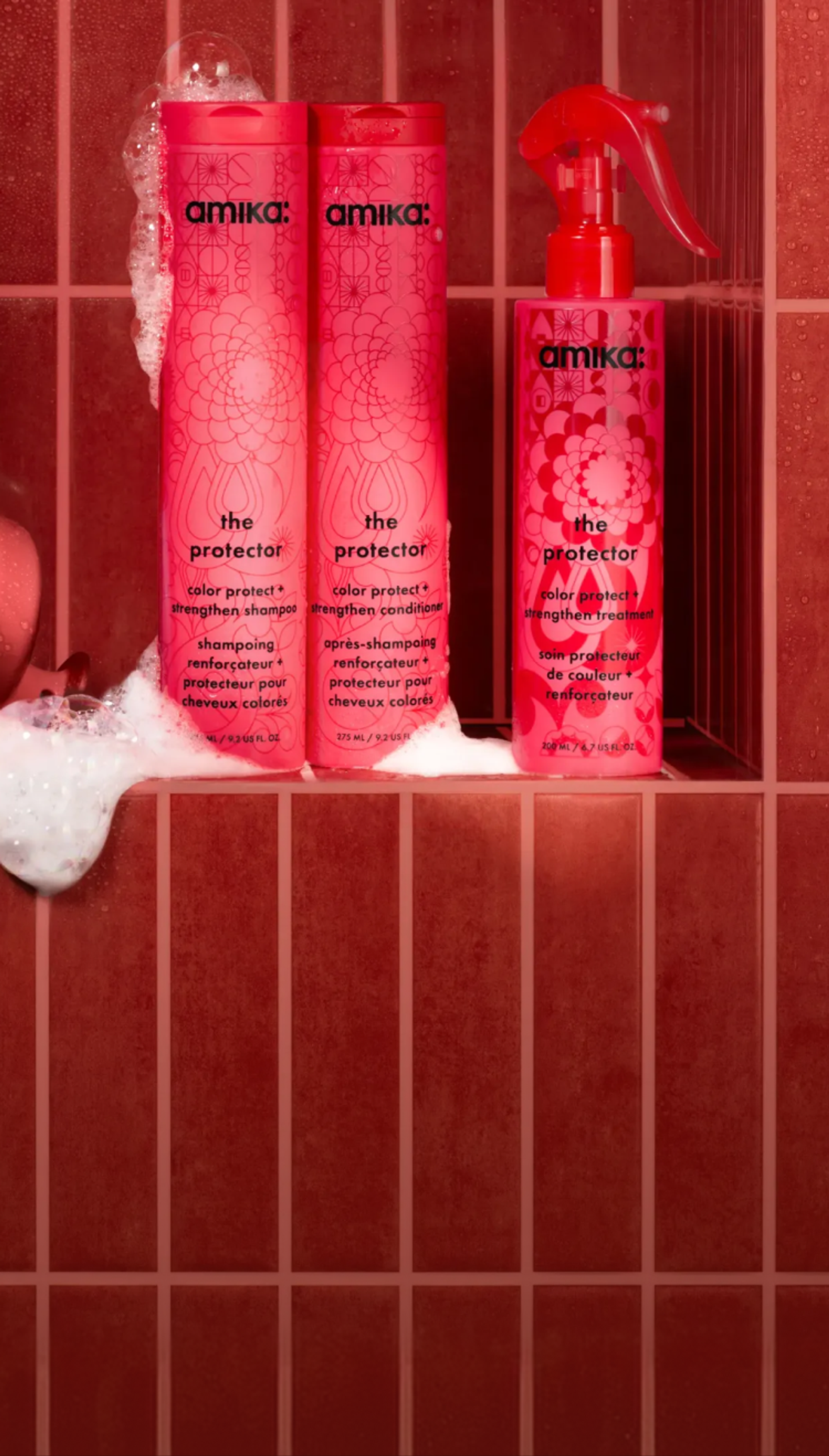 amika's the protector shampoo and conditioner and treatment bottles sit in a red tiled shower with a red scalp brush and suds