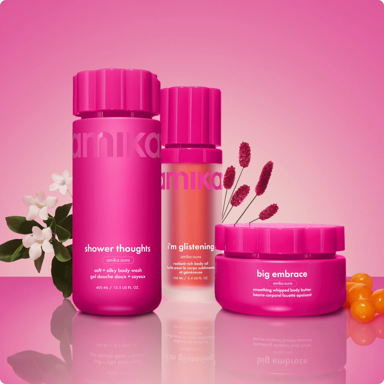 amika shower thoughts body wash, i'm glistening body oil, and big embrace body butter sit against a pink gradient background with flowers and sea buckthorn berries