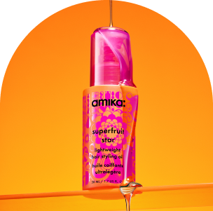 a pink and orange bottle of amika superfruit star lightweight hair styling oil sits on a clear shelf and the oil is being poured over and down the bottle