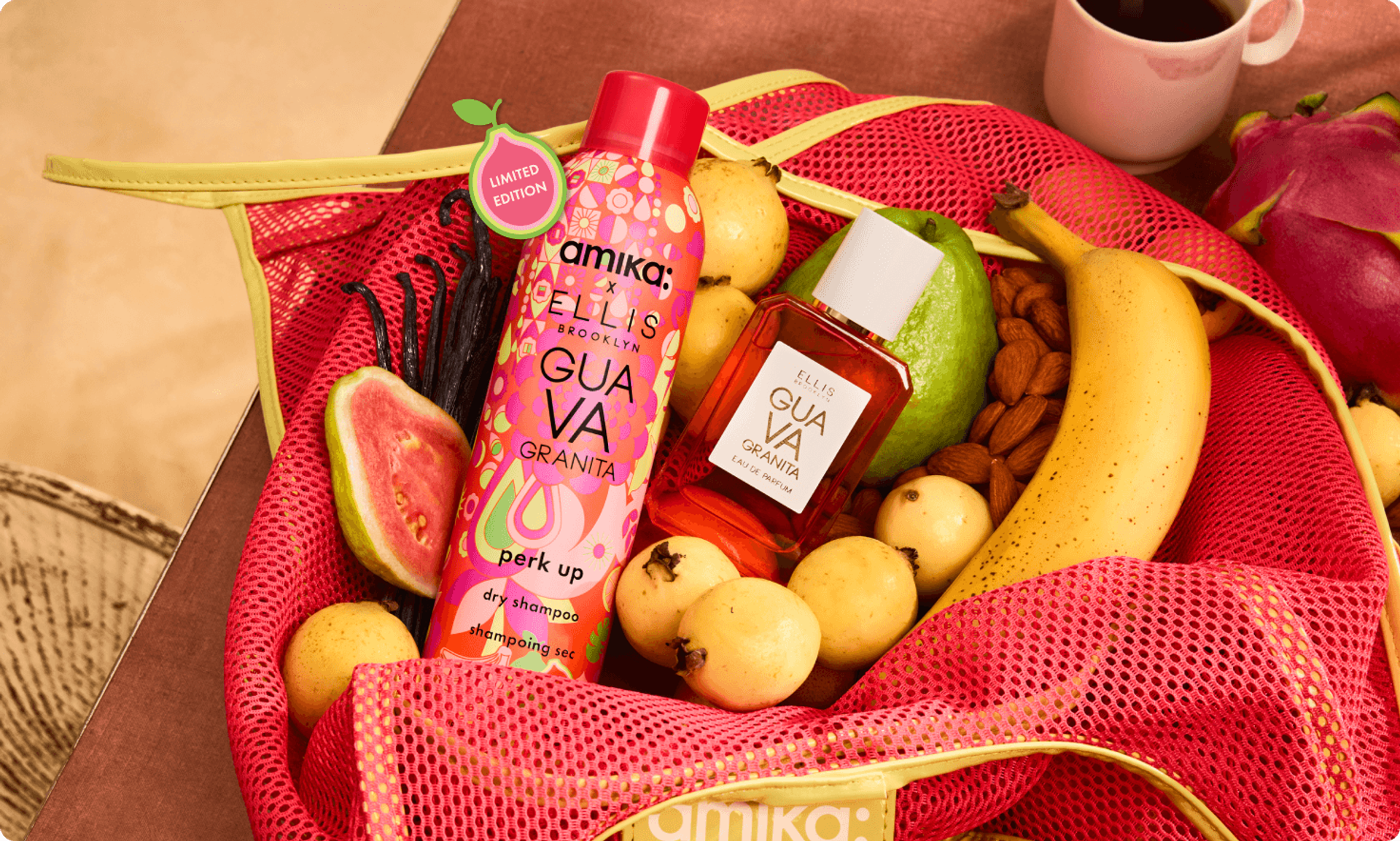 a bottle of amika x Ellis Brooklyn limited-edition perk up GUAVA GRANITA dry shampoo and a bottle of Ellis Brooklyn GUAVA GRANITA perfume sit in a pink mesh bag with fruit and almonds surrounding them