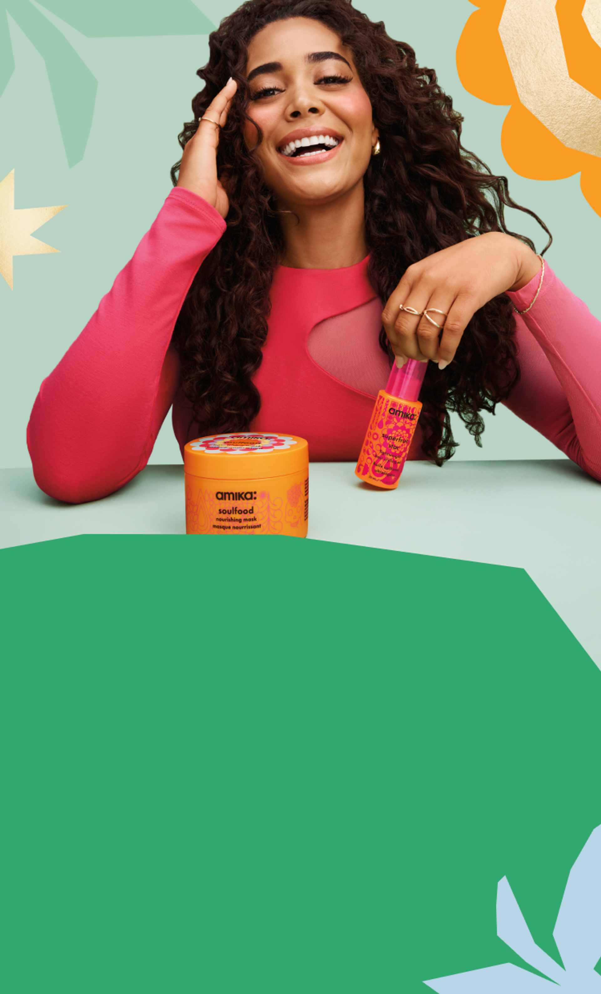 model with long, brown, curly hair smiles while holding a bottle of amika superfruit star hair oil. a jar of soulfood hair mask sits in the foreground and the background is light green with a darker green leaf shape in the middle and a flower or sun shape in the right hand corner