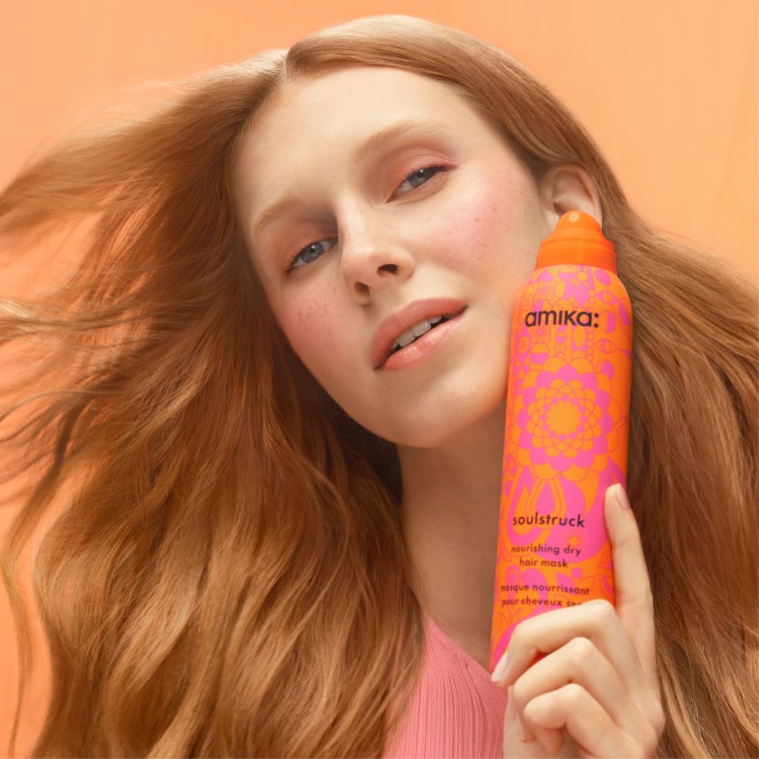 a model with soft, flowy reddish-blond hair holds a bottle of amika soulstruck spray in front of a light orange background