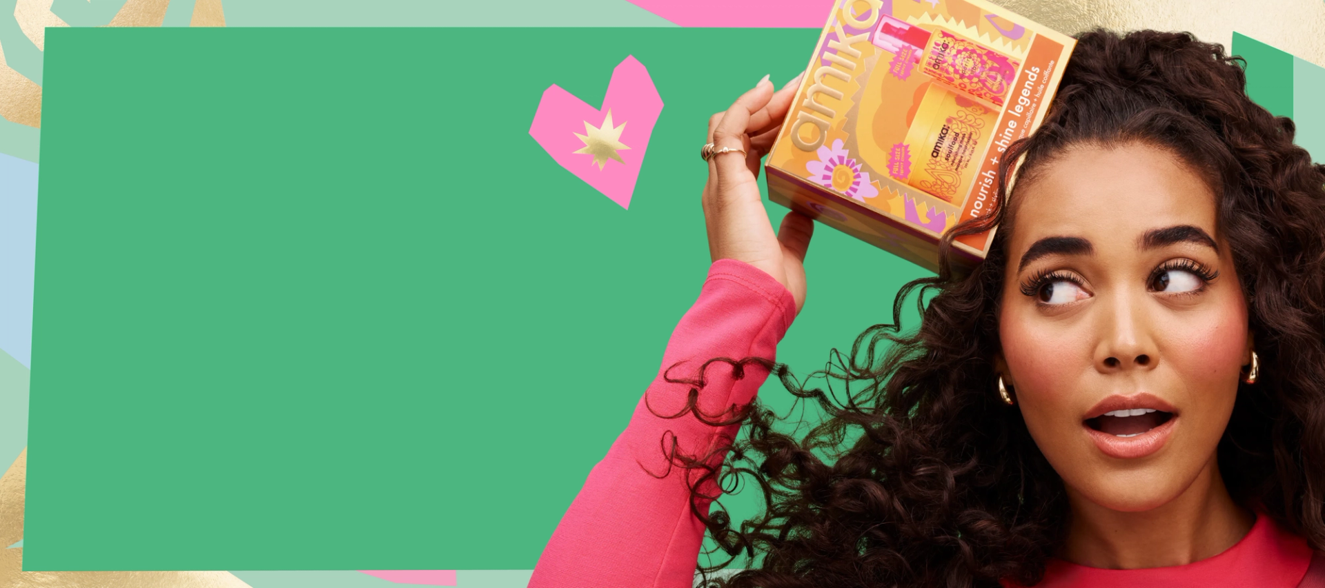 a model with long, brown, curly hair smiles while holding amika nourish + shine legends haircare gift set box. the background is green and there is a pink heart in the middle and a pastel border.