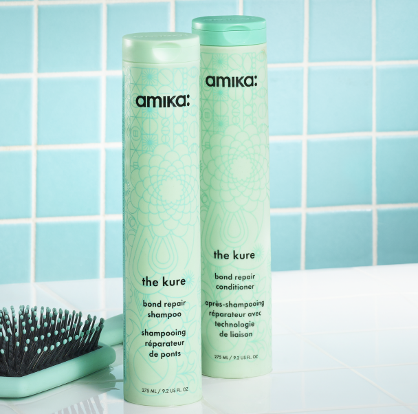 amika the kure strength repair shampoo and conditioner bottles sit on a white surface with a green brush next to them and in front of a blue-green tiled background