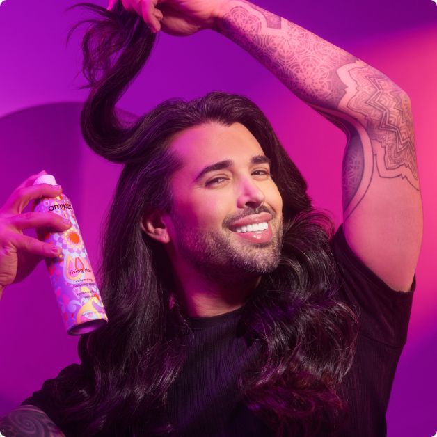 image of Eric Vaughn, a stylist for Amika, is smiling while holding a can of Amika's "Rising Star" volumizing spray in one hand and lifting a section of his long, voluminous hair with the other. Eric is wearing a black top and has visible tattoos on his arm. The background features a purple and pink gradient, enhancing the lively atmosphere. The "Rising Star" can is prominently displayed, showcasing its colorful, decorative packaging with intricate patterns.
