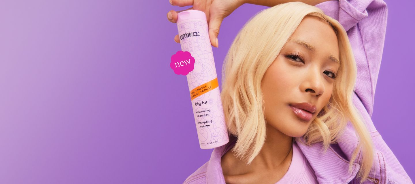 image of a model against a purple background. Model is posed holding the standard retail size of our NEW big hit volumizing shampoo (275ml / 9. 2 oz).