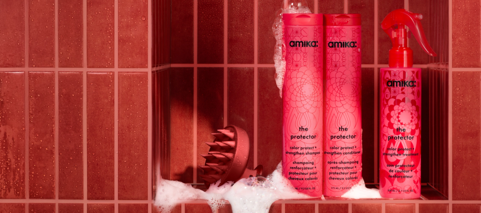 amika's the protector shampoo and conditioner and treatment bottles sit in a red tiled shower with a red scalp brush and suds