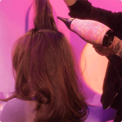 a GIF (moving image) of a hair stylist using an amika blow dryer to add volume to the root area of a model's long brown hair