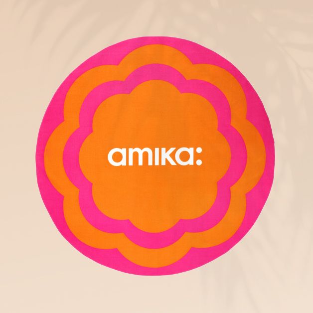 image of a pink and orange patterned circular towel with a white amika logo on a sand background with slight palm leaves shadows