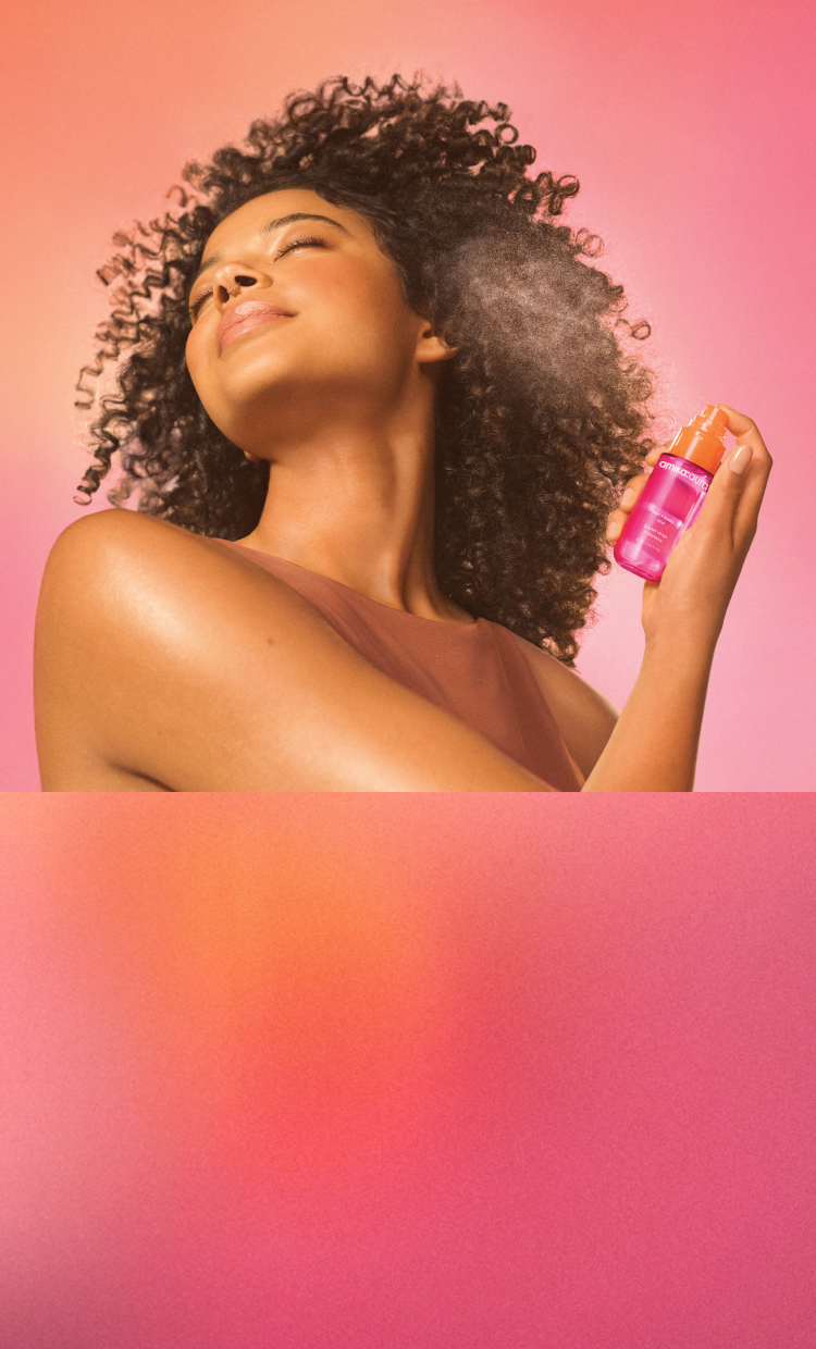 a model mists amika:aura hair and body mist onto their hair. model is set against a pink gradient background