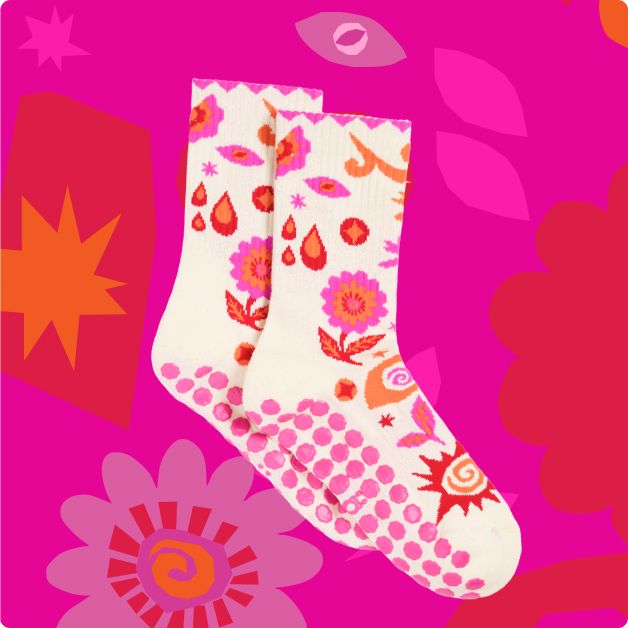 a pair of patterned socks with pink grips on the bottom against a pink, red, dark pink, and orange floral pattern.