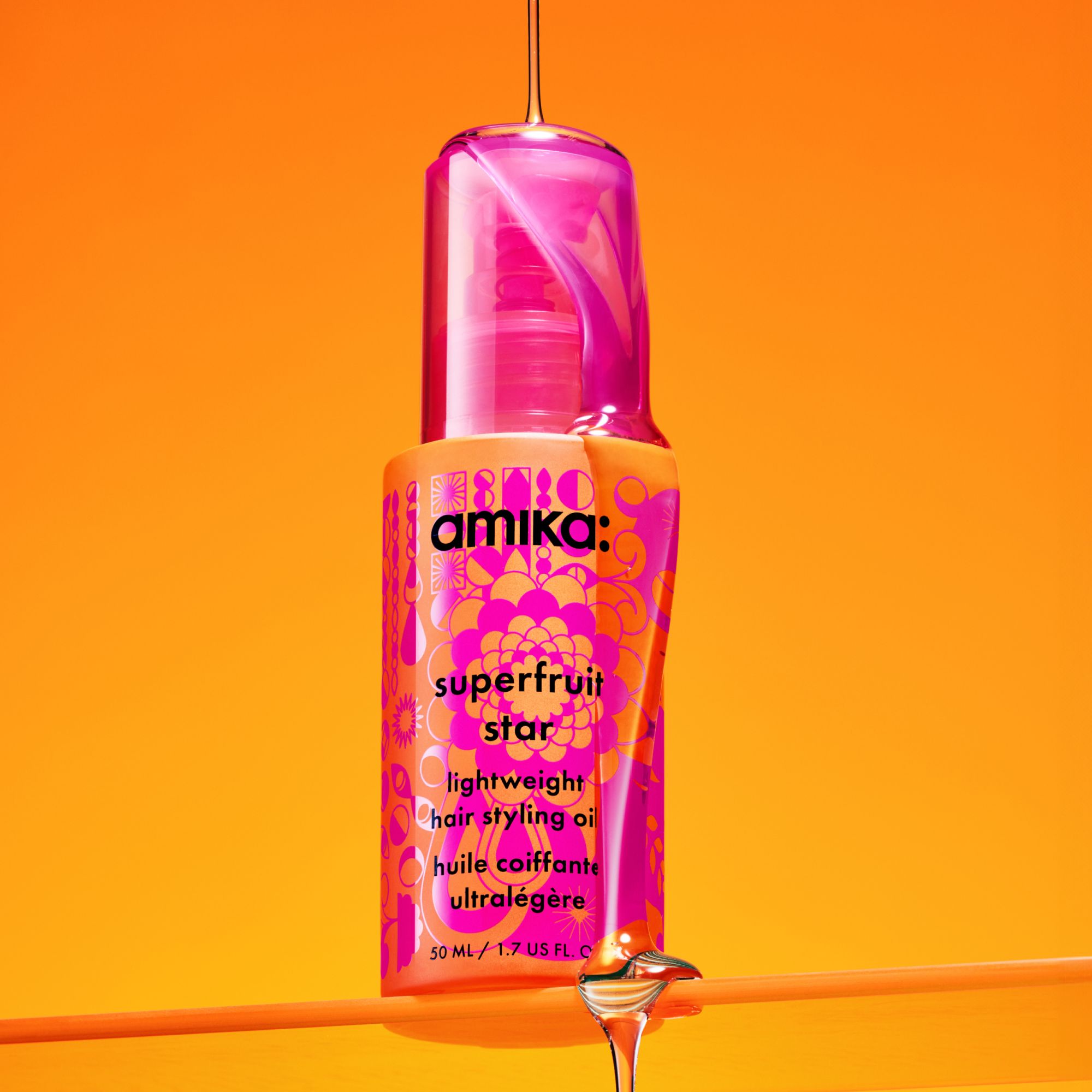 a pink and orange bottle of amika superfruit star lightweight hair styling oil sits on a clear shelf and the oil is being poured over and down the bottle