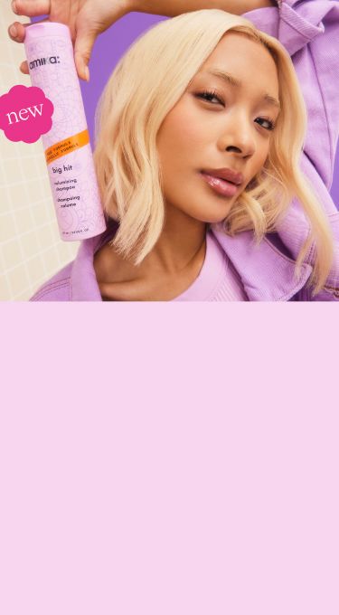 image of a model against a purple background. Model is posed holding the standard retail size of our NEW big hit volumizing shampoo (275ml / 9. 2 oz).