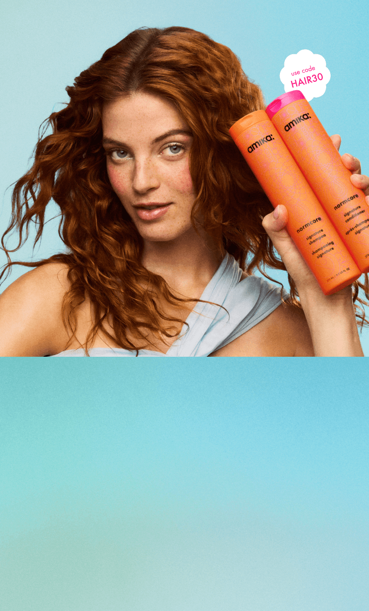 model with wavy red hair holds amika normcore shampoo + conditioner bottles against a light blue background