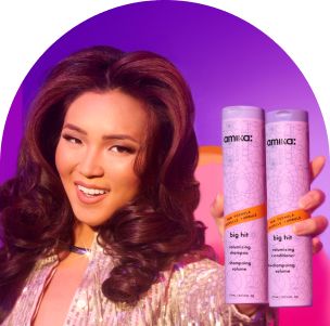 image of a smiling model with voluminous, styled hair holds two bottles of Amika's "Big Hit" volumizing shampoo and conditioner. The model is dressed in a shiny, metallic outfit, complementing the vibrant purple and pink gradient background. The "Big Hit" bottles are prominently displayed in the model's hand, showcasing their decorative packaging with intricate patterns. The overall image highlights the product's ability to create volume and the playful, energetic branding of Amika.