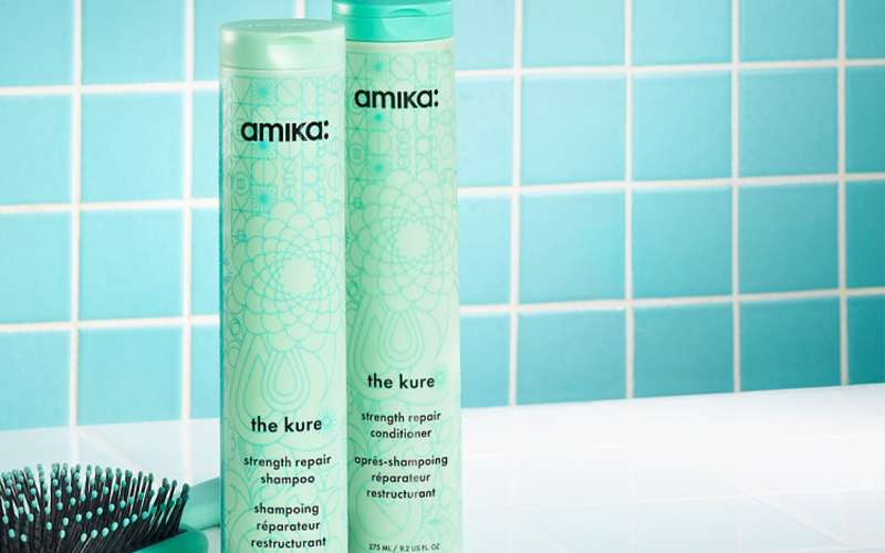 amika the kure strength repair shampoo and conditioner bottles sit on a white surface with a green brush next to them and in front of a blue-green tiled background