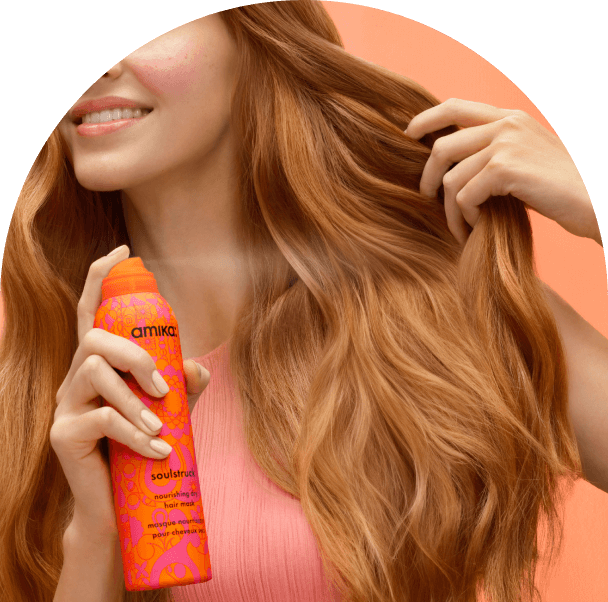 model spraying amika soulstruck dry mask in their hiar in front of a pinkish-orange background