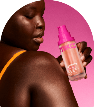 a model holds a bottle of amika i'm glistening body oil near their shoulder. There is a pink background