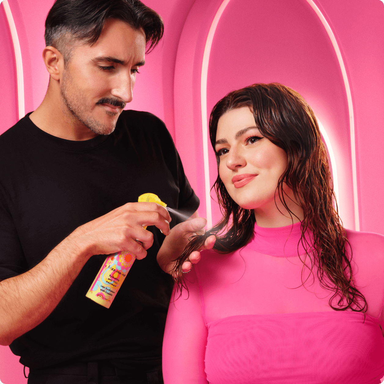 a stylist applies amika's frizz-me-not hydrating anti-frizz treatment spray to a person with damp, wavy hair in a pink salon environment