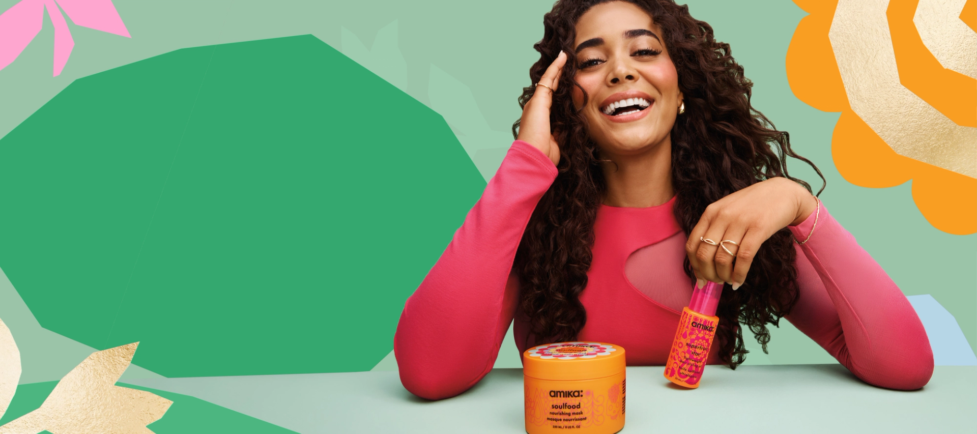 model with long, brown, curly hair smiles while holding a bottle of amika superfruit star hair oil. a jar of soulfood hair mask sits in the foreground and the background is light green with a darker green leaf shape in the middle and a flower or sun shape in the right hand corner