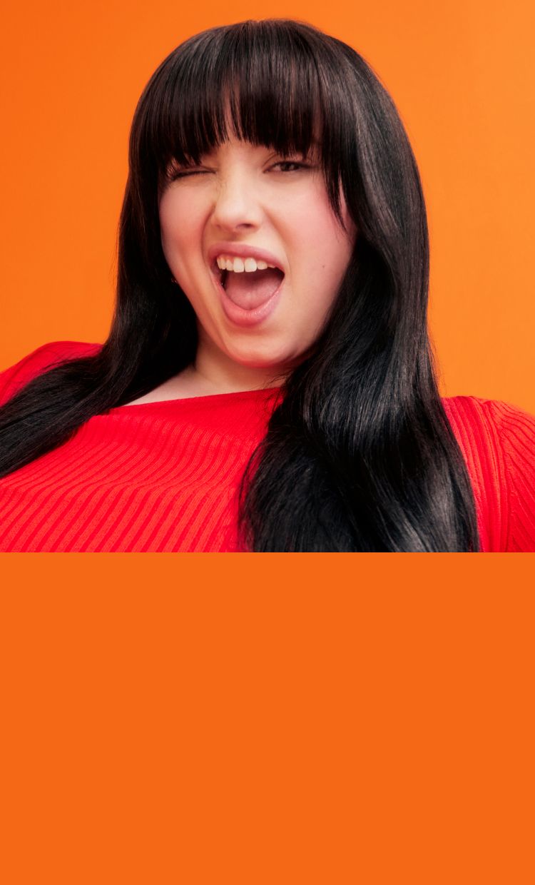 amika model with brunette, straight hair in front of an orange background and wearing an orange shirt