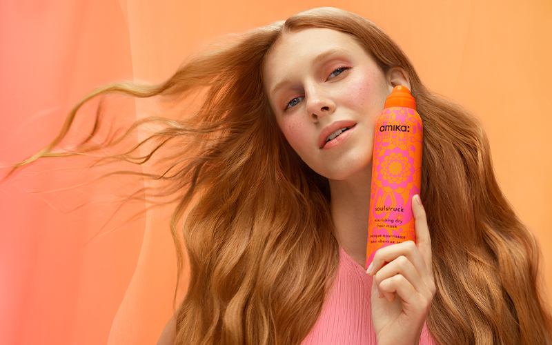 a model with soft, flowy reddish-blond hair holds a bottle of amika soulstruck spray in front of a light orange background