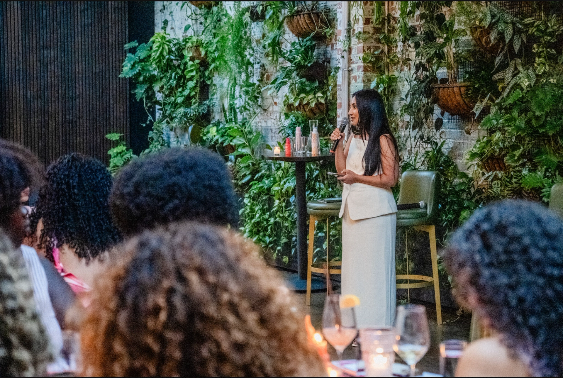 amika CMO Nilofer Vahora speaking to an audience in front of a wall with leaves and greenery