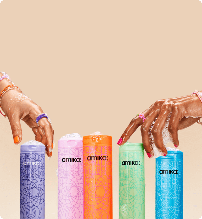 two hands with soapy lather on them reach for amika shampoo bottles against a cream background