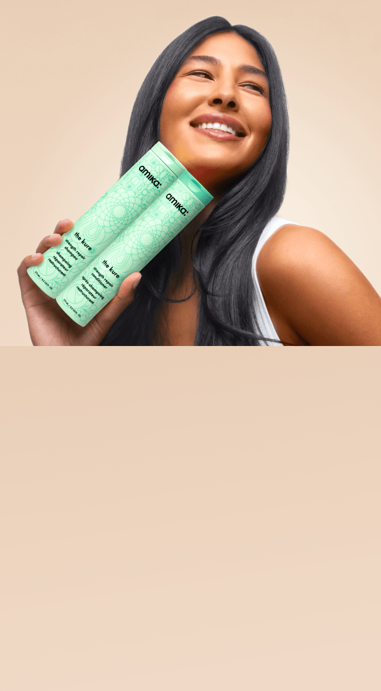 a model holds bottles of amika the kure shampoo and conditioner against a tan background