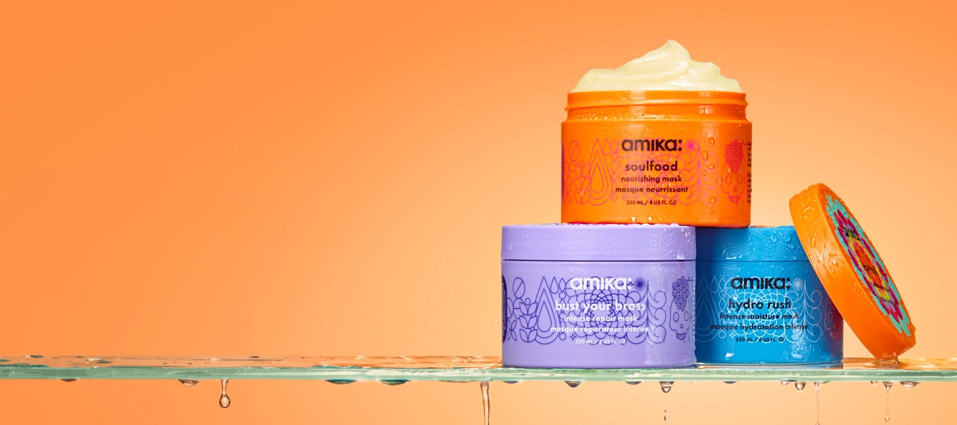 amika masks: hydro rush, soulfood, and bust your brass sit on a wet glass surface in front of an orange background