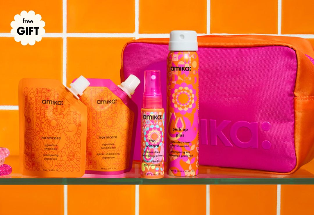 free gift with your custom amika on-the-go travel bundle. Travel pouches of shampoo + conditioner, a spray primer, a travel dry shampoo and a pink and orange cosmetic bag sit on a shelf against an orange tiled background