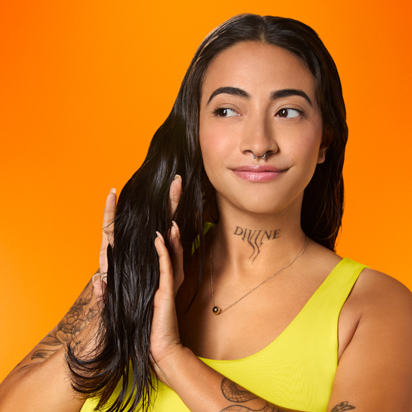 Showing dry hair application with superfruit star lightweight hair styling oil used on model Amber Perez shown in a bright yellow shirt, with dark hair, against an orange hued background