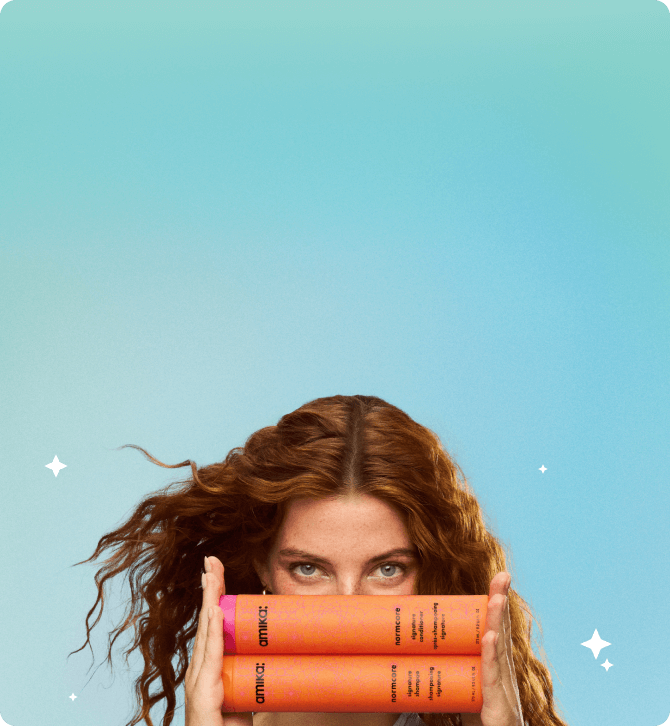 a model holds amika normcore shampoo and conditioner bottles horizontally while against a bluish background