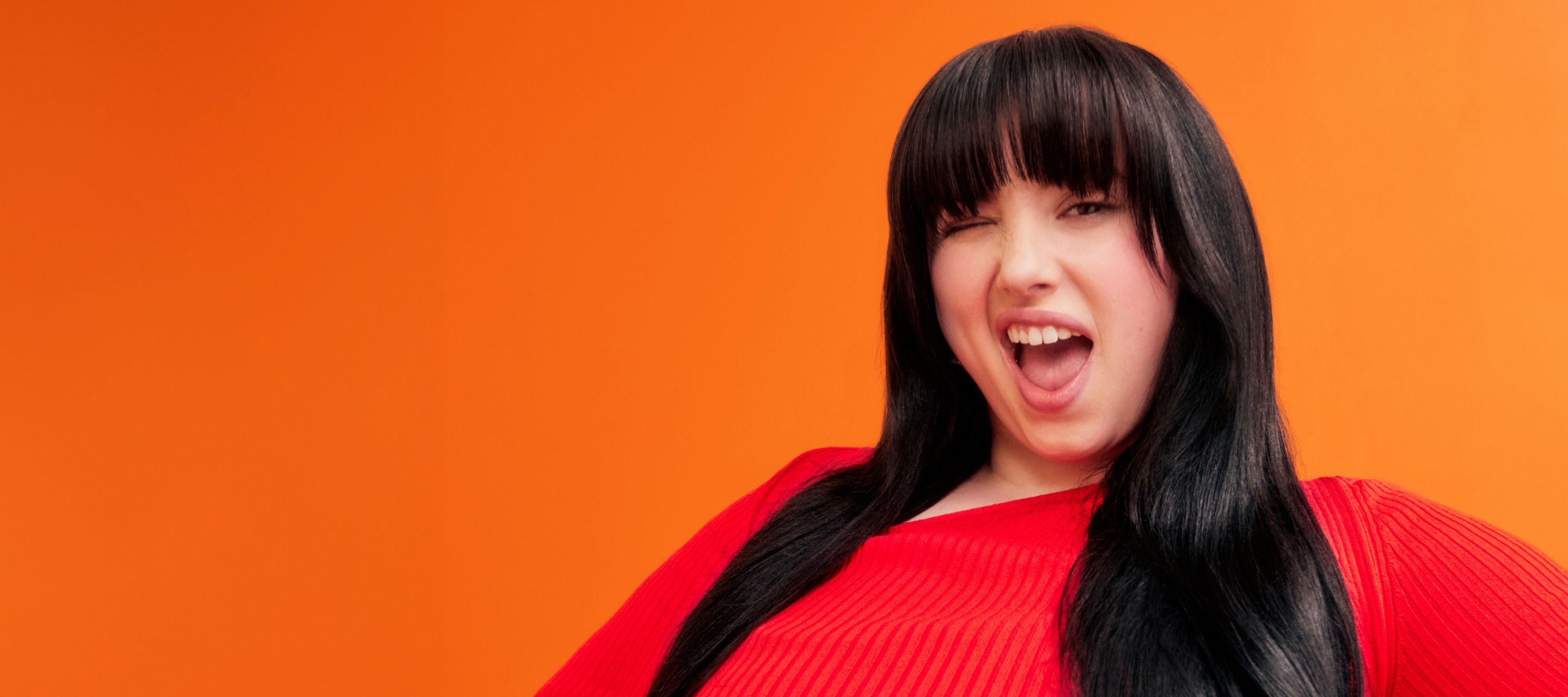 amika model with brunette, straight hair in front of an orange background and wearing an orange shirt