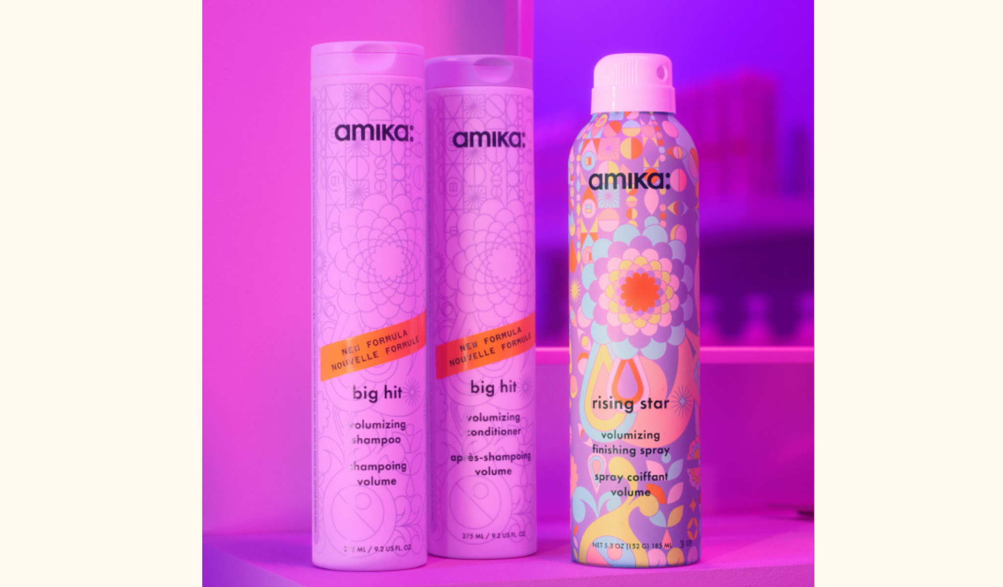 image of display of Amika's new volumizing hair care products set against a purple background. The image features three products: two tall bottles of "Big Hit" volumizing shampoo and conditioner on the left, and one aerosol can of "Rising Star" volumizing finishing spray on the right. Each product has decorative packaging with intricate patterns and vibrant colors. The background features a pink and purple gradient, emphasizing the playful and stylish branding of Amika. The image showcases the volumizing benefits of the "Big Hit" shampoo and conditioner, as well as the "Rising Star" finishing spray.