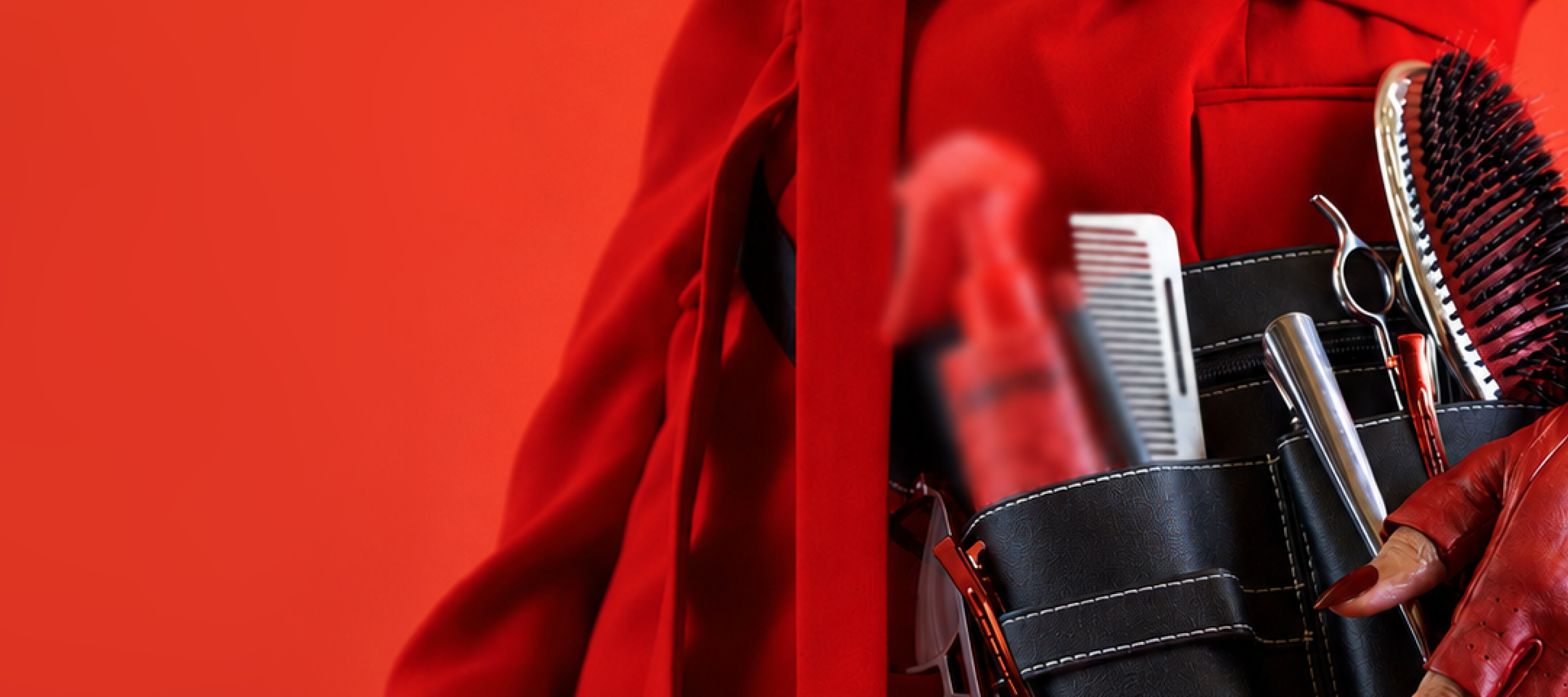 a closeup of a hairstylist's tool belt with scissors, comb, brush, and a blurred-out product. the person is wearing all red.