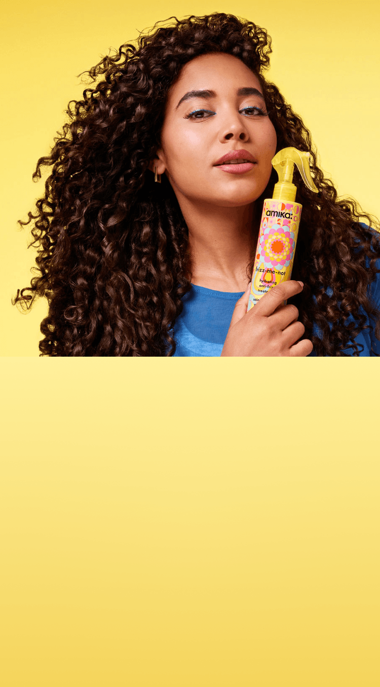 a model holds a bottle of amika frizz-me-not treatment against a yellow background.