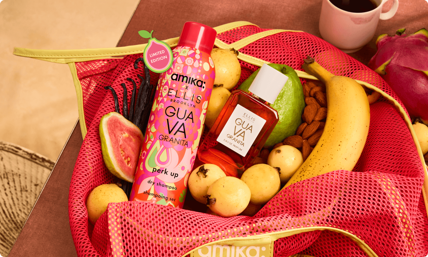 a bottle of amika x Ellis Brooklyn limited-edition perk up GUAVA GRANITA dry shampoo and a bottle of Ellis Brooklyn GUAVA GRANITA perfume sit in a pink mesh bag with fruit and almonds surrounding them