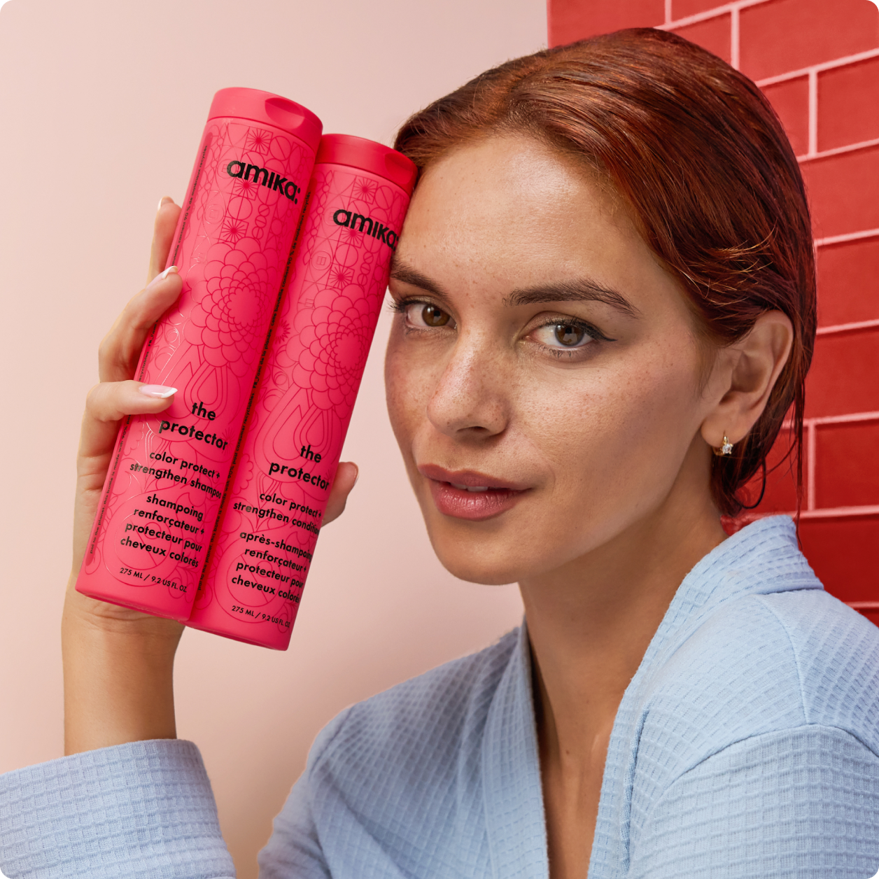 a model with red hair holds bottles of amika the protector shampoo and conditioner