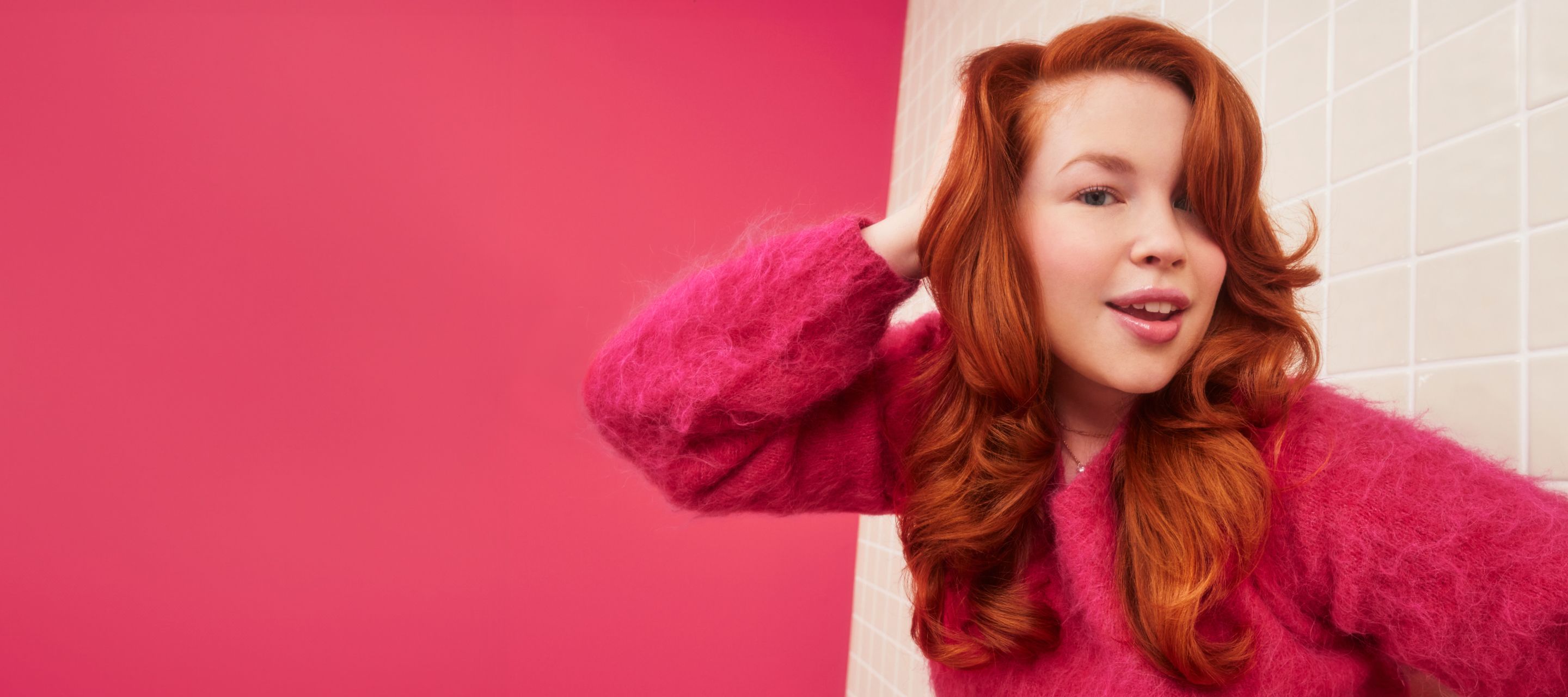 model with red, wavy hair wearing a pink sweater in front of a cream tile background