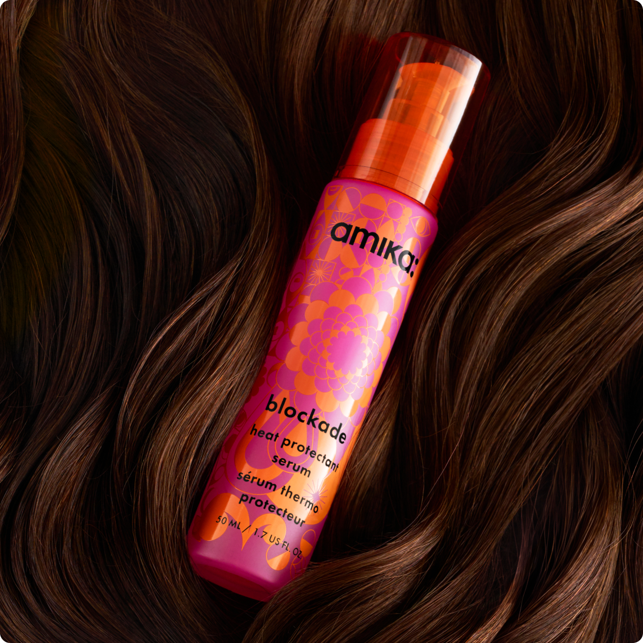 a pink and orange bottle of amika blockade heat protectant serum lies nestled in brown, wavy hair