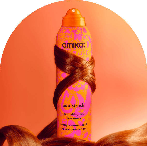 a tress of reddish brown hair wraps around amika soulstruck spray. the bottle sits in front of a pinkish-orange background