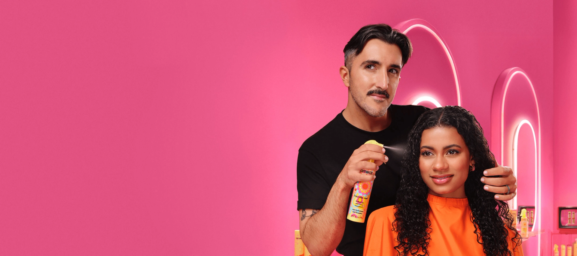 a stylist applies amika's frizz-me-not hydrating anti-frizz treatment spray to a person with curly hair in a pink salon environment