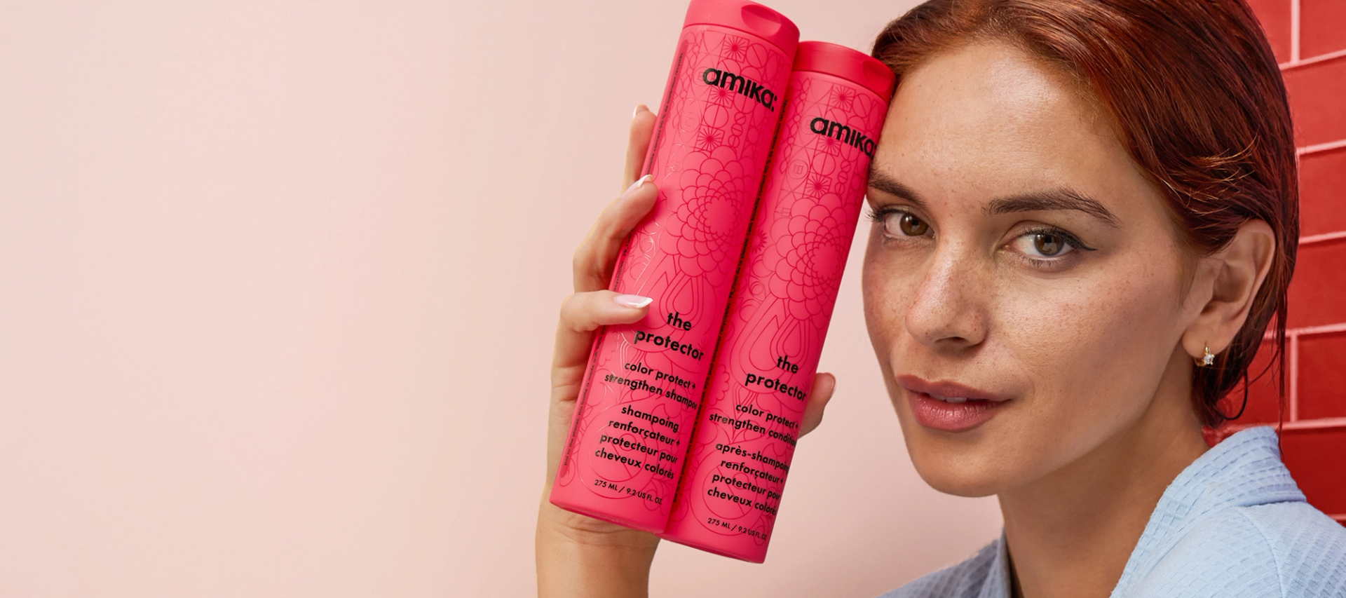 a model with red hair holds bottles of amika the protector shampoo and conditioner