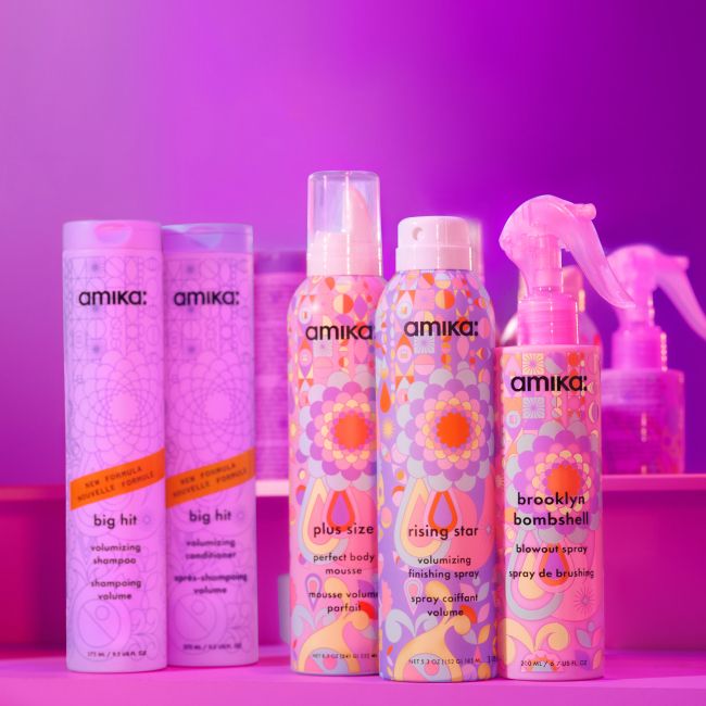 A vibrant display of Amika's volumizing hair care products set against a purple background. The image features five products: two tall bottles of "Big Hit" volumizing shampoo and conditioner on the left, two aerosol cans of "Plus Size" and "Rising Star" volumizing finishing spray in the center, and a spray bottle of "Brooklyn Bombshell" blowout spray on the right. Each product has colorful, decorative packaging with intricate patterns. The background includes additional spray bottles, emphasizing the lively and playful branding of Amika's hair care line.