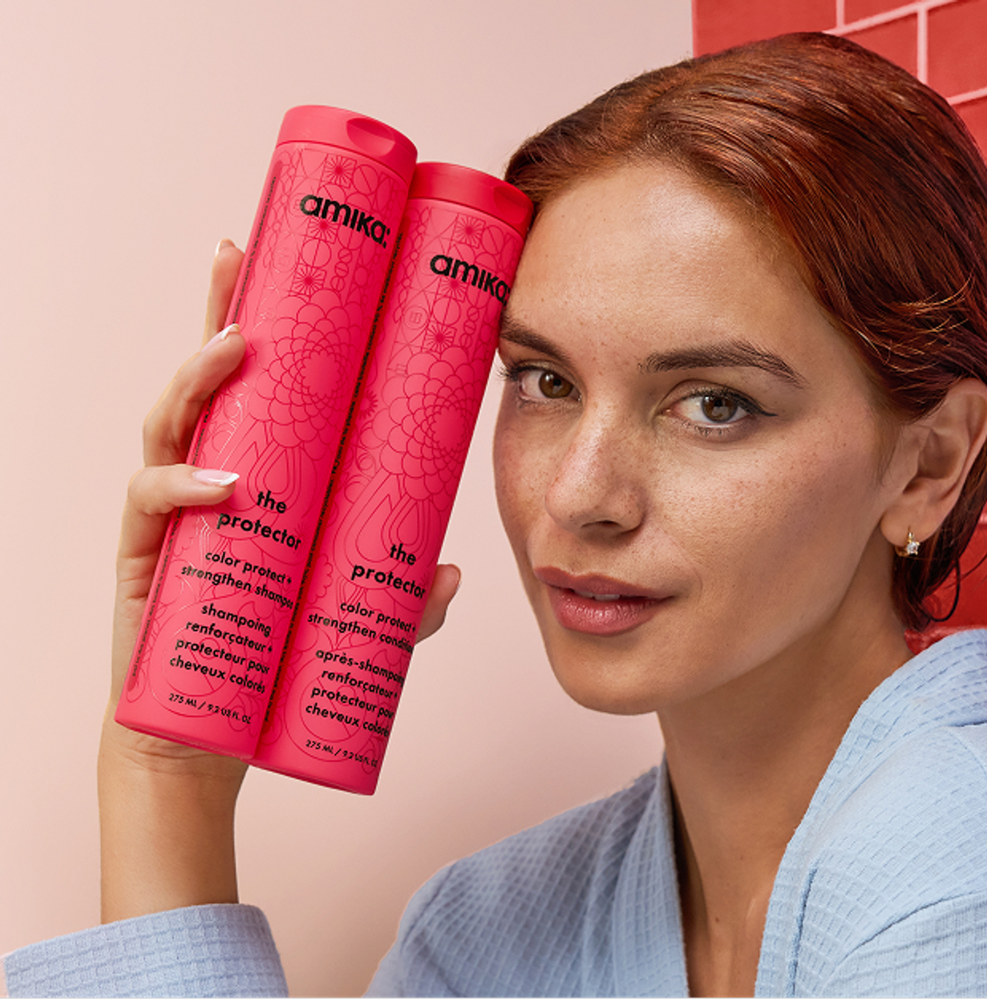 a model with red hair holds bottles of amika the protector shampoo and conditioner