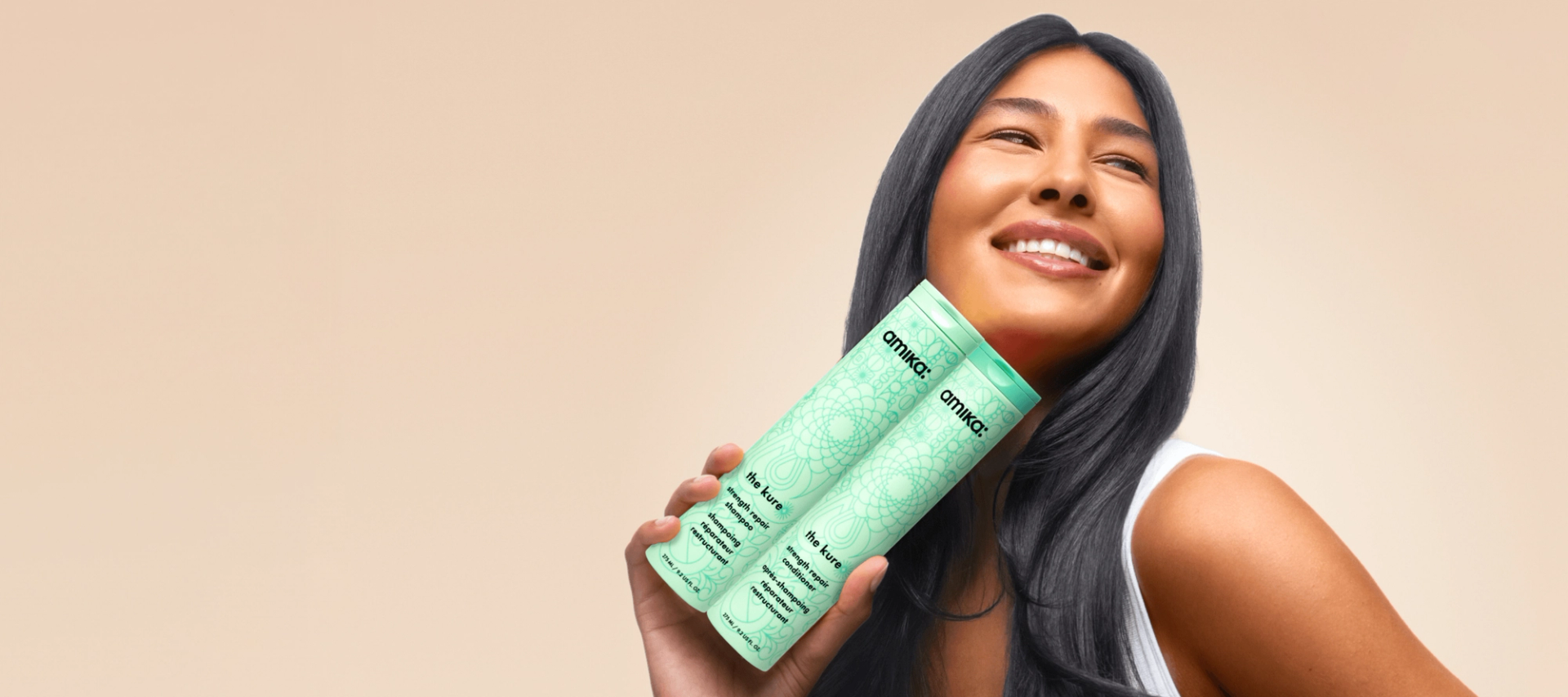 a model holds bottles of amika the kure shampoo and conditioner against a tan background