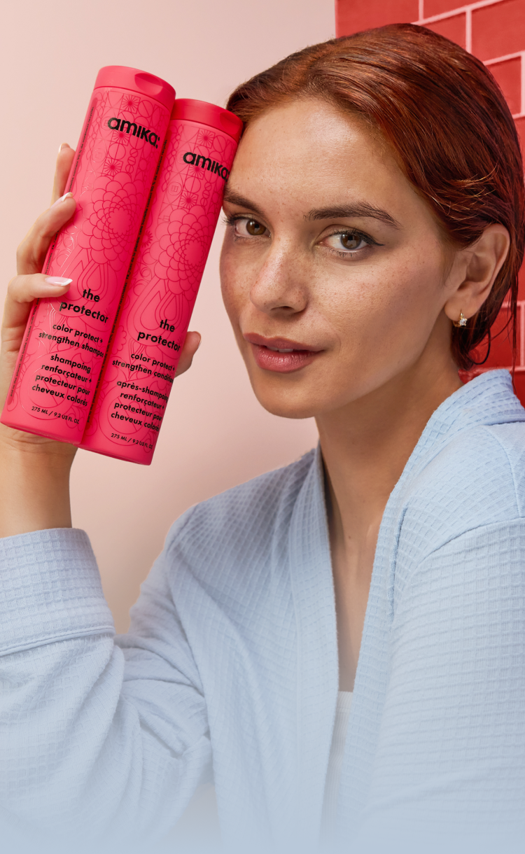 a model with red hair holds bottles of amika the protector shampoo and conditioner